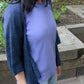 A person wearing a light blue shirt and the Knitbot Wispy Cardi by Never Not Knitting stands outdoors in a garden setting. Tall green plants are visible in the background, along with raised garden beds made of wood. The person's face is not visible in the image.