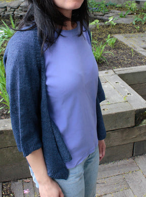 A person wearing a light blue shirt and the Knitbot Wispy Cardi by Never Not Knitting stands outdoors in a garden setting. Tall green plants are visible in the background, along with raised garden beds made of wood. The person's face is not visible in the image.