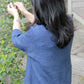 A person with long dark hair, wearing the Knitbot Wispy Cardi by Never Not Knitting and light blue jeans, is seen from behind as they reach up to pick or inspect something on a leafy green plant with small pink flowers. The scene is set outdoors near a house with wooden siding and a deck.