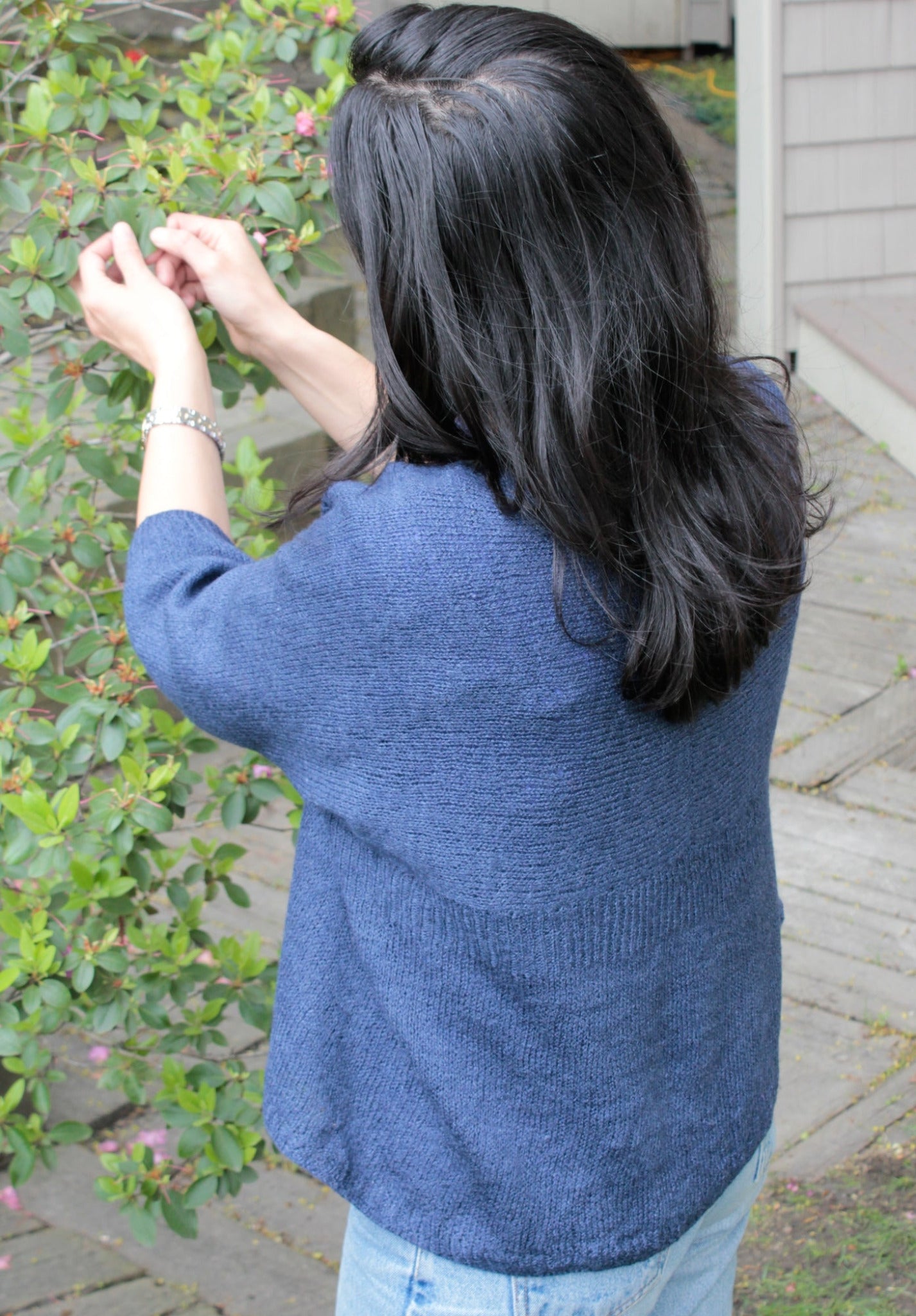 A person with long dark hair, wearing the Knitbot Wispy Cardi by Never Not Knitting and light blue jeans, is seen from behind as they reach up to pick or inspect something on a leafy green plant with small pink flowers. The scene is set outdoors near a house with wooden siding and a deck.