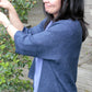 A woman with long dark hair and a light complexion is standing outdoors, smiling as she reaches up to touch leaves on a bush. She is wearing the blue Knitbot Wispy Cardi by Never Not Knitting over a purple top and a wristwatch. A wooden deck and part of a building are visible in the background.