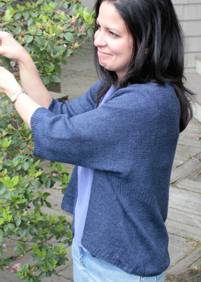 A woman with long dark hair and a light complexion is standing outdoors, smiling as she reaches up to touch leaves on a bush. She is wearing the blue Knitbot Wispy Cardi by Never Not Knitting over a purple top and a wristwatch. A wooden deck and part of a building are visible in the background.