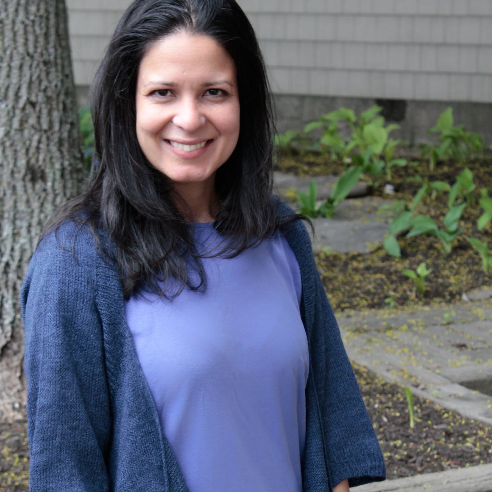 A woman with long, dark hair smiling while standing outside, wearing a purple shirt and the Knitbot Wispy Cardi in dark blue by Never Not Knitting. A tree trunk and green plants are visible in the background.