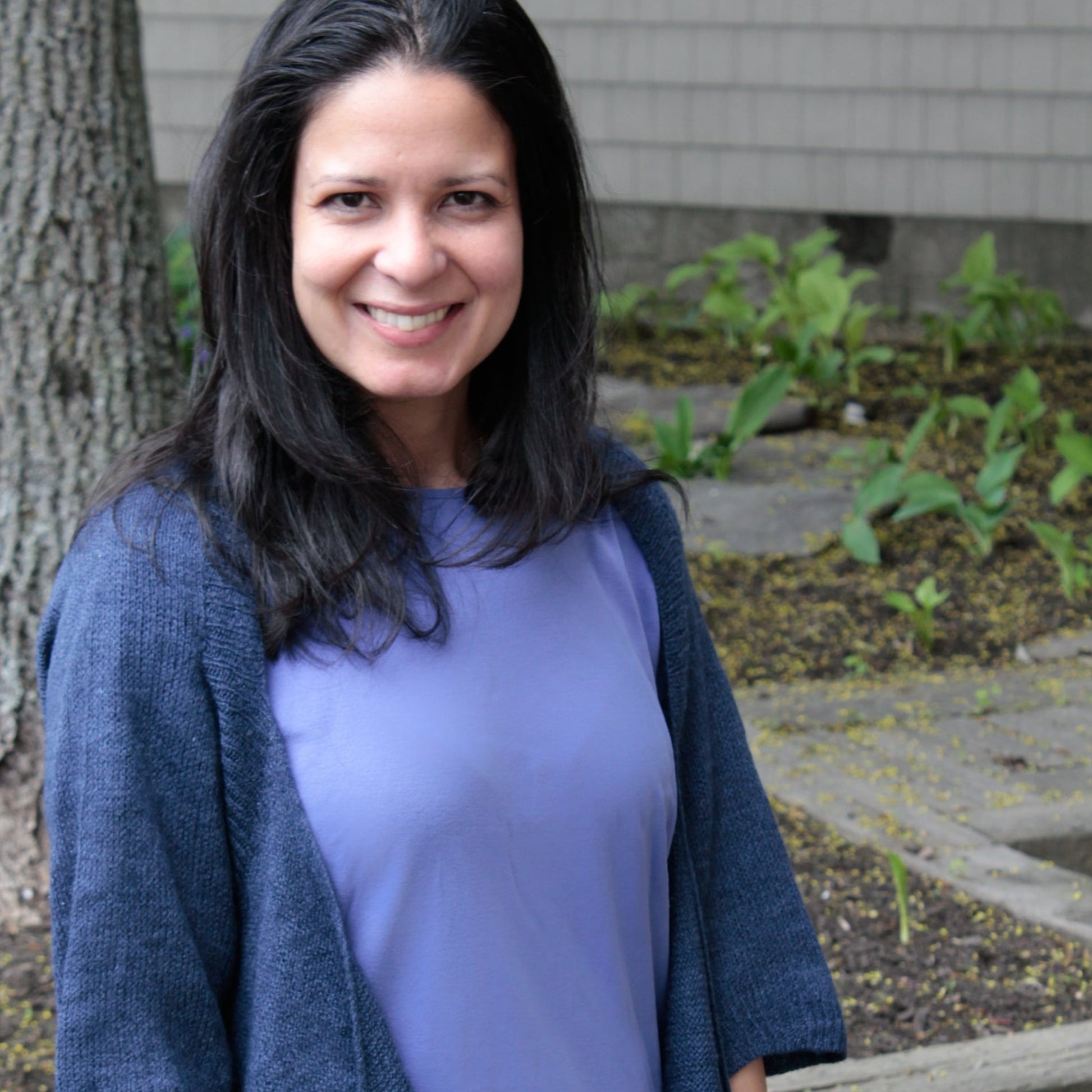 A woman with long dark hair and a broad smile stands outdoors near a tree. She is wearing a purple top paired with the Knitbot Wispy Cardi by Never Not Knitting in dark blue. The ground around the tree has some fresh green plants, and the background consists of a wooden structure, hinting at nearby construction work.