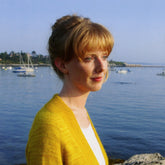 A woman with red hair and a Knitbot Featherweight Cardigan by Never Not Knitting stands by the water, gazing thoughtfully into the distance. Several sailboats float on the calm water in the background, and a shoreline with trees and houses is visible under a clear, blue sky.