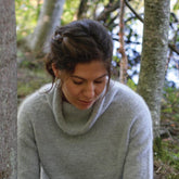 A woman with brown hair, gently tied back, is wearing a soft Knitbot Lightweight Pullover from Never Not Knitting. She is looking down and smiling slightly. She is outdoors, surrounded by trees and a softly blurred forest background as sunlight filters through the leaves.