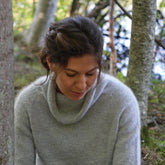 A woman with brown hair is sitting outdoors among trees, looking down with a peaceful expression. Dressed in the Knitbot Lightweight Pullover from Never Not Knitting, made from soft dk weight yarn and featuring a light gray hue, she has a braid in her hair. The background is filled with green foliage and the hint of a body of water, creating a serene scene.