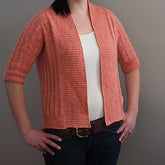 A person is wearing the Knitbot Spring Ribbed Cardigan from Never Not Knitting in a fresh coral color over a white tank top, paired with blue jeans. Their hands are placed on their hips, standing against a plain, light gray background. This versatile three-season cardigan features ribbed patterns and three-quarter-length sleeves, making it an excellent addition to any wardrobe.