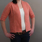 A person with dark hair stands against a plain backdrop, wearing the vibrant Knitbot Spring Ribbed Cardigan in a fresh coral hue by Never Not Knitting over a white top. Their hands rest on their hips, and they pair the outfit with dark jeans and a belt. The photo captures the upper body, highlighting this versatile wardrobe addition's textured details.
