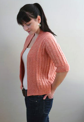 A woman with her hair tied in a ponytail, wearing a white top and the Knitbot Spring Ribbed Cardigan in peach with three-quarter sleeves from Never Not Knitting, poses sideways facing right. She has her hands in the pockets of her dark jeans. She is smiling gently against a plain light background. This cardigan is the perfect wardrobe addition for any season.