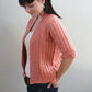 A person with dark hair, tied back in a ponytail, stands in profile against a plain background. They are wearing the peach-colored Knitbot Spring Ribbed Cardigan from Never Not Knitting over a white top and blue jeans. With their hands in their pockets, they look down.