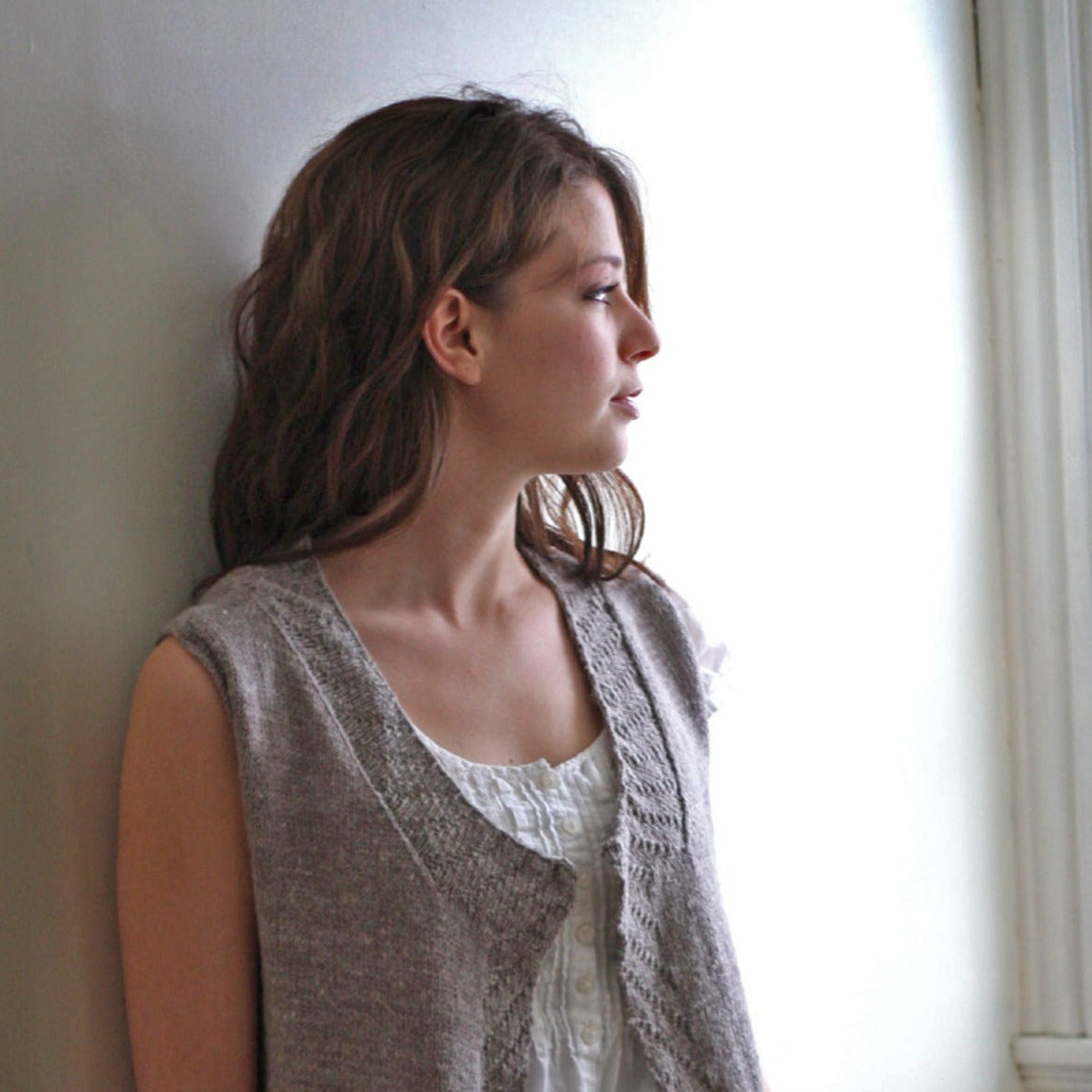 A woman with long brown hair is standing against a light-colored wall and gazing out of a window. She wears a loose-fit white blouse and the Knitbot Sycamore Vest by Never Not Knitting, featuring a sleeveless design and ribbed texture. The natural light from the window softly illuminates her profile.