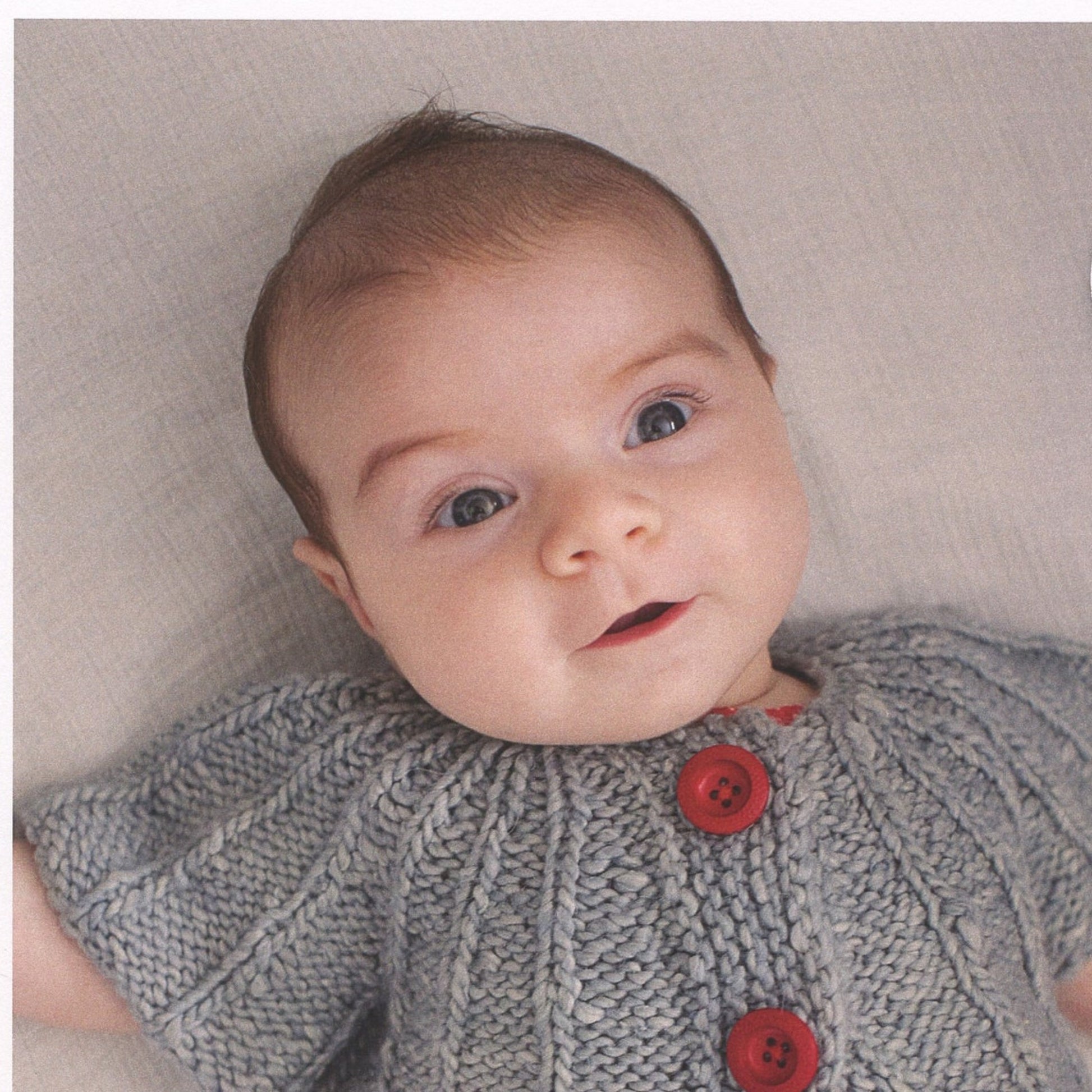 A baby with light skin, wide open eyes, and short brown hair is lying on a soft surface. The baby is wearing the Knitbot Yoked Cardigan from Never Not Knitting, which features a knitted gray ribbed design with large red buttons. The baby appears calm and content.