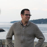 A person with glasses is standing outdoors in front of a scenic waterfront view, smiling as they look off into the distance. They are wearing a gray Knitbot Zip-Up Cardigan by Never Not Knitting over a checkered shirt. The background includes water, distant land, and sailboats.