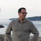 A person with glasses is standing outdoors in front of a scenic waterfront view, smiling as they look off into the distance. They are wearing a gray Knitbot Zip-Up Cardigan by Never Not Knitting over a checkered shirt. The background includes water, distant land, and sailboats.