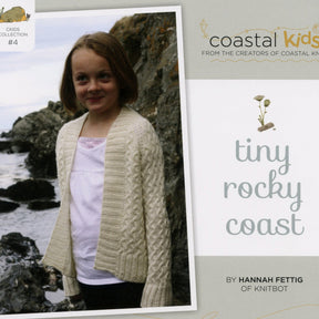 A young girl stands on a rocky coastline wearing a cream-colored knit cardigan over a white shirt. The image is the cover for "Coastal Kids Tiny Rocky Coast Cardigan" from Never Not Knitting, with floral illustrations and text indicating it’s part of the Coastal Kids collection by Hannah Fettig of Knitbot, featuring the Rocky Coast Cardigan in children's sizes.