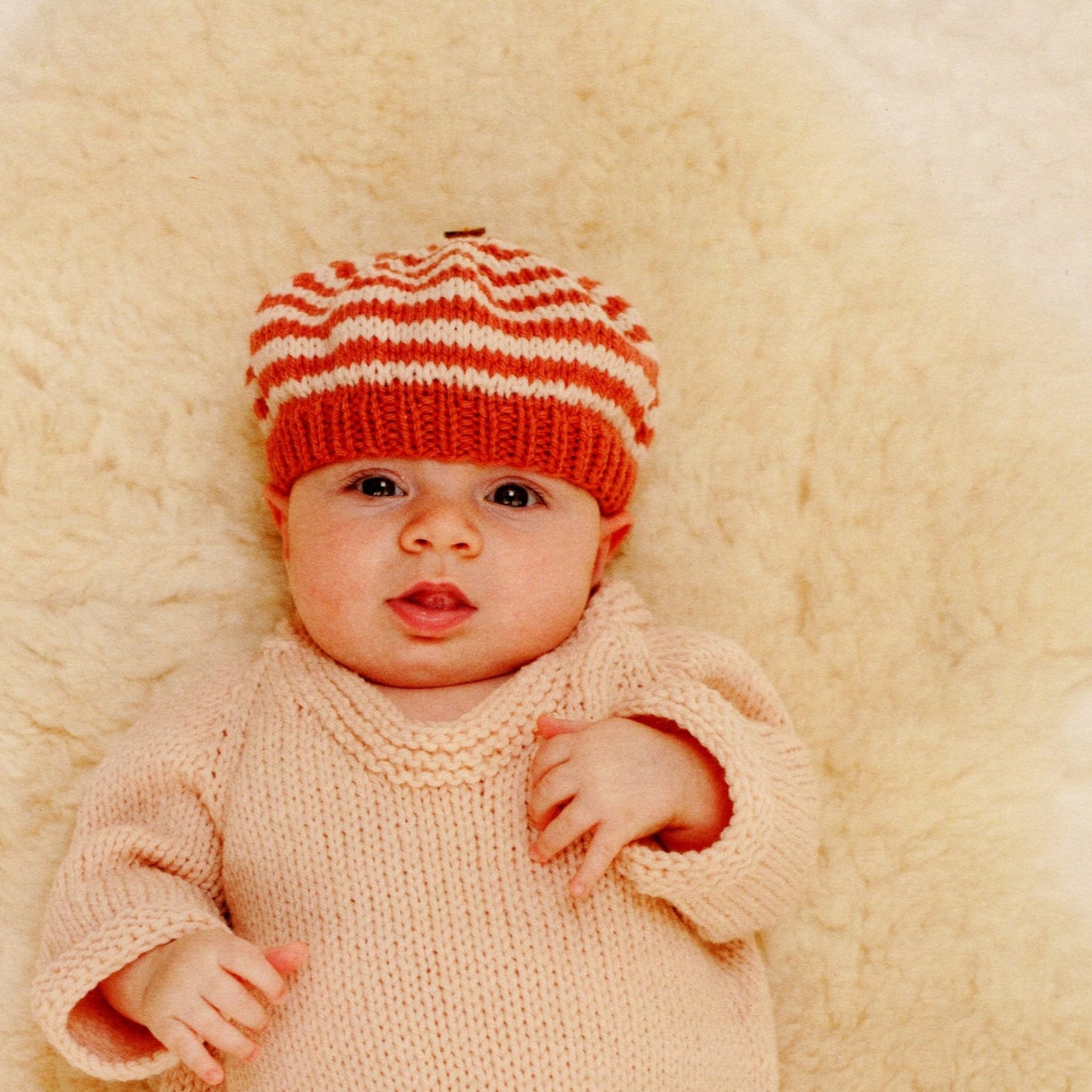 A baby wearing Mabel's Closet Autumn Frock & Stripe Hat from Never Not Knitting lies on a soft, fuzzy surface, looking up with a neutral expression. The background is a light cream color.