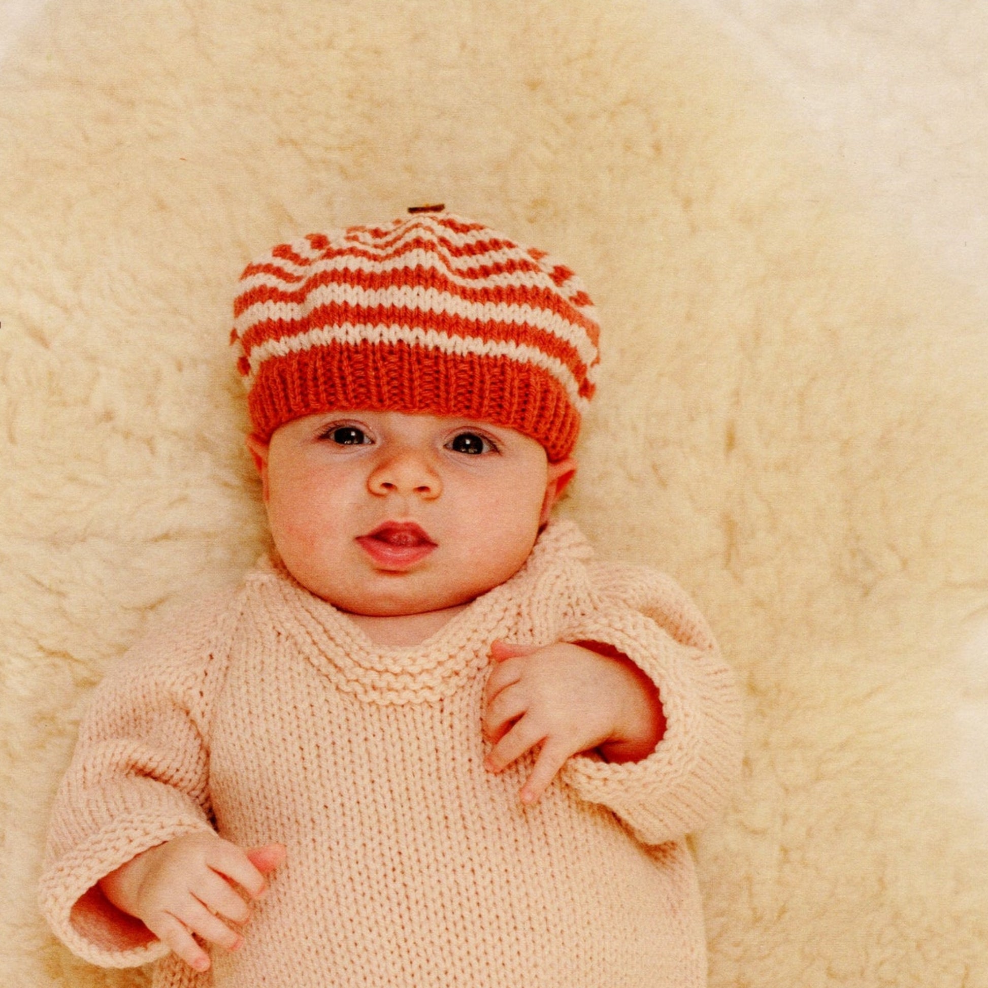 A little one lies on a soft, cream-colored fuzzy blanket, wearing Mabel's Closet Autumn Frock & Stripe Hat by Never Not Knitting. The baby has wide eyes and is looking directly at the camera with a slightly open mouth.