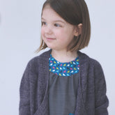 A little girl with shoulder-length brown hair is smiling and looking to the side. She is wearing the Never Not Knitting Knitbot Bitty Breezy Cardigan, a sophisticated dark gray open front cardigan, over a gray dress featuring a blue, multi-colored pattern near the neckline and a matching pocket. The background is plain white.