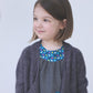 A little girl with shoulder-length brown hair is smiling and looking to the side. She is wearing the Never Not Knitting Knitbot Bitty Breezy Cardigan, a sophisticated dark gray open front cardigan, over a gray dress featuring a blue, multi-colored pattern near the neckline and a matching pocket. The background is plain white.