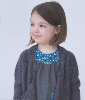 A little girl with shoulder-length brown hair is smiling and looking to the side. She is wearing the Never Not Knitting Knitbot Bitty Breezy Cardigan, a sophisticated dark gray open front cardigan, over a gray dress featuring a blue, multi-colored pattern near the neckline and a matching pocket. The background is plain white.