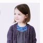 A sophisticated little one with shoulder-length brown hair smiles while looking to her left. She is wearing a blue dress with a colorful patterned neckline and the Knitbot Bitty Breezy Cardigan in purple from Never Not Knitting. The background is plain white.