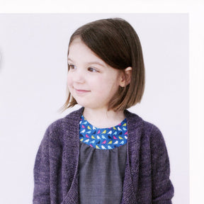 A sophisticated little one with shoulder-length brown hair smiles while looking to her left. She is wearing a blue dress with a colorful patterned neckline and the Knitbot Bitty Breezy Cardigan in purple from Never Not Knitting. The background is plain white.