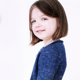 A young child with short brown hair, wearing a blue long-sleeved Knitbot Fledgling Featherweight Cardigan by Never Not Knitting, is smiling and looking slightly off to the side. The background is a plain white, giving a clean and simple appearance to the image—reminiscent of the top-down simplicity for which Knitbot designs are known.