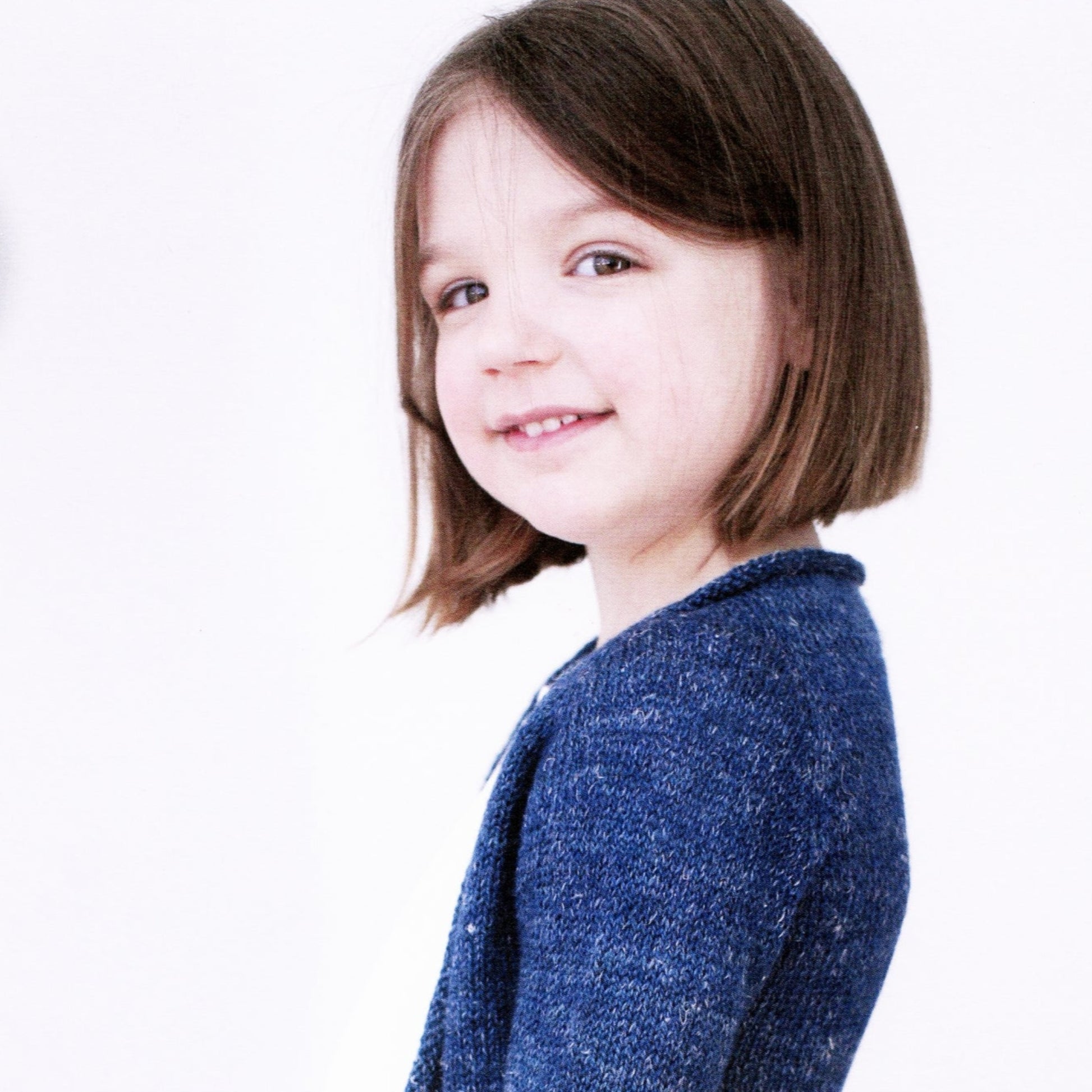 A young girl with shoulder-length brown hair is smiling while facing slightly to the right. She is wearing a blue, long-sleeve Knitbot Fledgling Featherweight Cardigan from Never Not Knitting, showcasing the simple top-down cardigan style. The background is plain white.