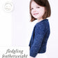 A young girl with shoulder-length brown hair is standing against a white background, smiling softly. She is wearing a Knitbot Fledgling Featherweight Cardigan by Never Not Knitting over a white shirt. The text on the image reads "fledgling featherweight," showcasing this delicate design by Hannah Fettig.