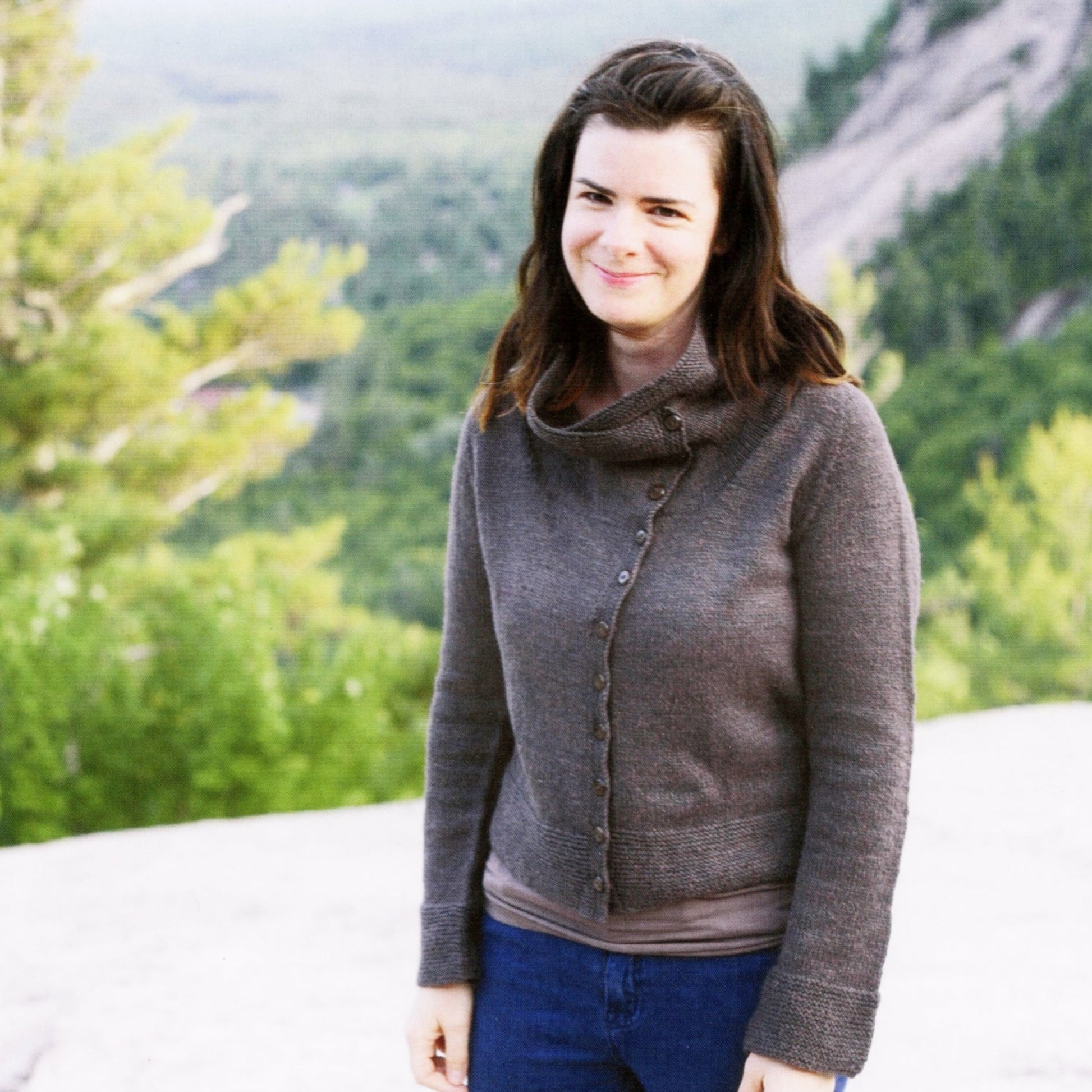 A woman with long dark hair is standing outdoors, smiling, wearing the Knitbot Moto Jacket by Never Not Knitting paired with blue jeans. The background showcases a blurred scenic view of trees and a rock formation under a cloudy sky.