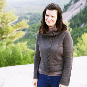 A woman with long dark hair is standing outdoors, smiling, wearing the Knitbot Moto Jacket by Never Not Knitting paired with blue jeans. The background showcases a blurred scenic view of trees and a rock formation under a cloudy sky.