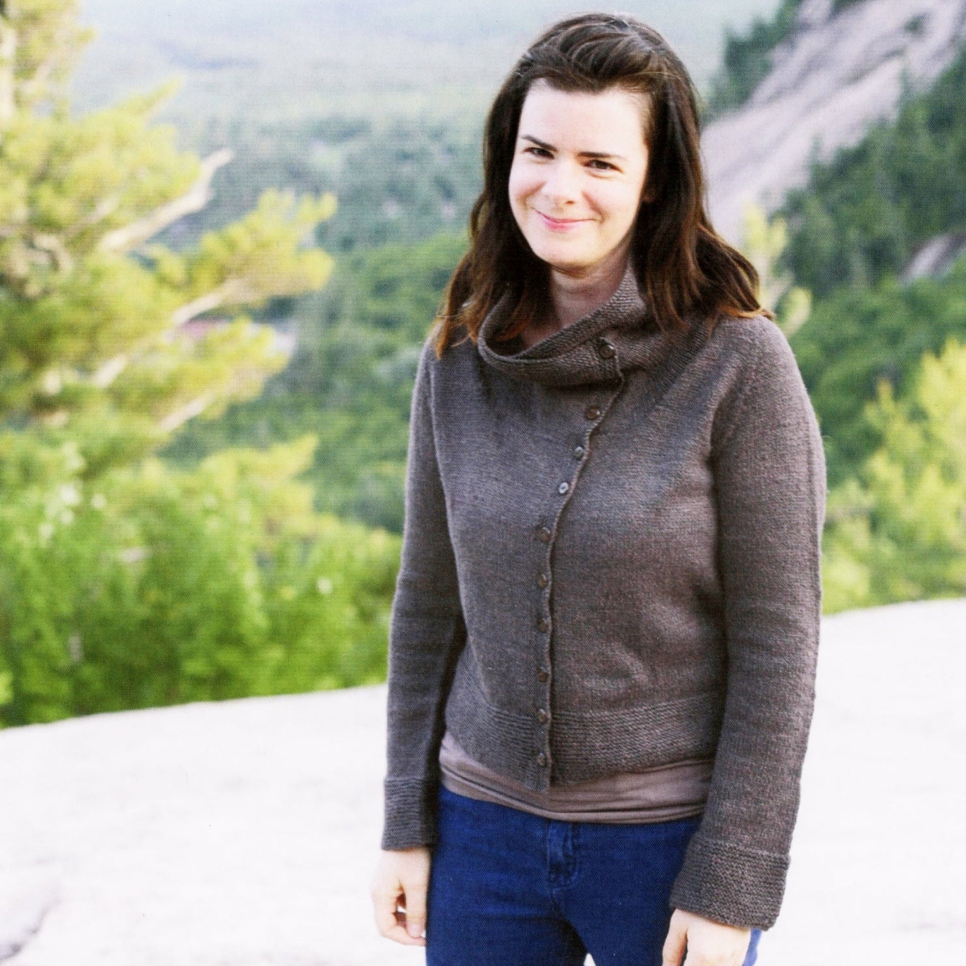 A woman with long brown hair wearing the Knitbot Moto Jacket by Never Not Knitting and blue jeans stands smiling outdoors. The background features blurred green foliage and a rocky landscape under a cloudy sky, perfectly showcasing her impeccable jacket styling.