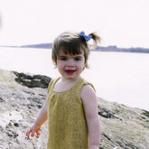 A young child with a blue bow in their hair smiles at the camera while standing on a rocky shore by a body of water. The child is wearing the sleeveless, yellow Knitbot Sweet Pleat girls top from Never Not Knitting. The background shows a calm body of water and distant tree line.