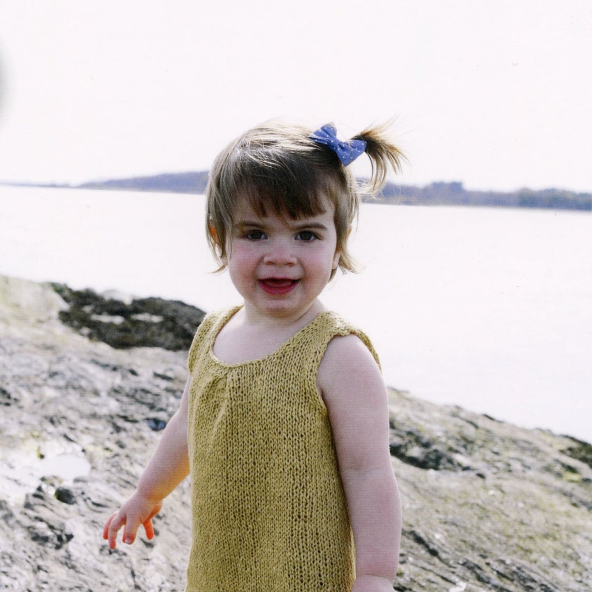 A young child with a blue bow in their hair smiles at the camera while standing on a rocky shore by a body of water. The child is wearing the sleeveless, yellow Knitbot Sweet Pleat girls top from Never Not Knitting. The background shows a calm body of water and distant tree line.