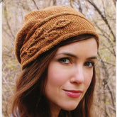 A woman with long brown hair is wearing the Sprig Cloche, a knitted hat by Never Not Knitting that features an asymmetrical brim and a leaf-like pattern. She is outdoors, against a backdrop of bare, leafless trees. With a slight smile, she gazes directly at the camera.