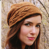 A woman with long brown hair is wearing a Sprig Cloche from Never Not Knitting. She is outdoors, set against a backdrop of autumn trees. The textured brown knit hat, knit flat, showcases an asymmetrical brim and a braided pattern running horizontally around it. She maintains a neutral expression on her face.