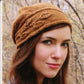 A woman with long brown hair is wearing a Sprig Cloche from Never Not Knitting. She is outdoors, set against a backdrop of autumn trees. The textured brown knit hat, knit flat, showcases an asymmetrical brim and a braided pattern running horizontally around it. She maintains a neutral expression on her face.