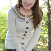 A woman with long brown hair is smiling and standing outdoors in front of leafy green trees. She is wearing the "Adult Playful Stripes" cardigan by Never Not Knitting, which features a colorful striped yoke and brown buttons, paired with dark blue jeans. She appears to be enjoying the natural surroundings.
