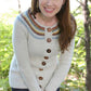 A woman with long brown hair is smiling and standing outdoors in front of leafy green trees. She is wearing the "Adult Playful Stripes" cardigan by Never Not Knitting, which features a colorful striped yoke and brown buttons, paired with dark blue jeans. She appears to be enjoying the natural surroundings.
