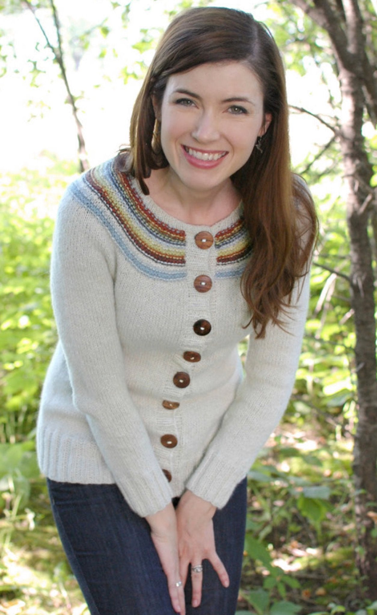 A woman with long brown hair is smiling and standing outdoors in front of leafy green trees. She is wearing the "Adult Playful Stripes" cardigan by Never Not Knitting, which features a colorful striped yoke and brown buttons, paired with dark blue jeans. She appears to be enjoying the natural surroundings.
