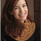 A woman with long red hair smiles warmly at the camera. She is wearing a Cream and Sugar Cowl from Never Not Knitting, featuring dramatic cables over a black top. The background showcases a brick wall with soft lighting, creating a cozy atmosphere.