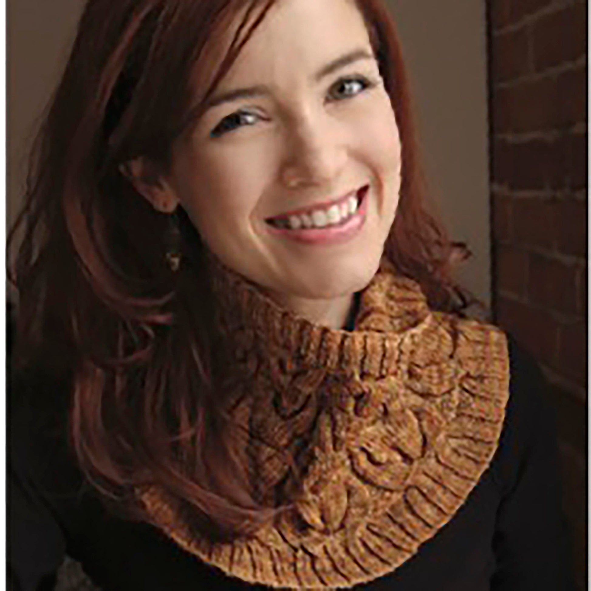 A woman with long red hair smiles warmly at the camera. She is wearing a Cream and Sugar Cowl from Never Not Knitting, featuring dramatic cables over a black top. The background showcases a brick wall with soft lighting, creating a cozy atmosphere.