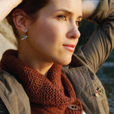 A person with short hair gazes thoughtfully into the distance. They wear a casual jacket and a cozy Shawl Collared Cowl, download by Never Not Knitting, crafted from bulky weight yarn. The background includes a blurry stone wall and water, with lighting suggesting it’s either sunset or early evening.