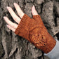 A hand wearing an Oak Grove mitt from Never Not Knitting, adorned with an oak leaf motif in a warm orange-brown hue, rests against the rough bark of a tree. This knitted fingerless mitten partially covers the wrist and leaves the fingertips exposed, blending perfectly with the texture of the tree bark in the background.