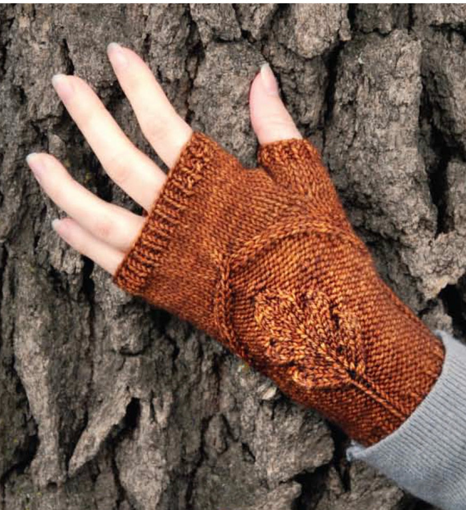 A hand wearing an Oak Grove mitt from Never Not Knitting, adorned with an oak leaf motif in a warm orange-brown hue, rests against the rough bark of a tree. This knitted fingerless mitten partially covers the wrist and leaves the fingertips exposed, blending perfectly with the texture of the tree bark in the background.