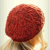 A person with long, blonde hair is shown from the back, wearing a beautifully knit Autumn Vines Beret by Never Not Knitting. The red beret, featuring a detailed leafy design, appears warm and cozy, covering the top and back of the head and ears.