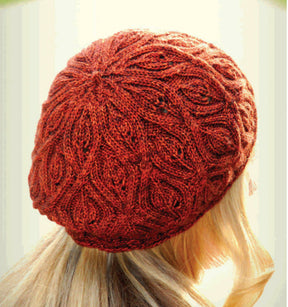 A person with long, blonde hair is shown from the back, wearing a beautifully knit Autumn Vines Beret by Never Not Knitting. The red beret, featuring a detailed leafy design, appears warm and cozy, covering the top and back of the head and ears.