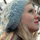 A woman with long blonde hair is gazing upwards. She is wearing the Cloudy Day Beret from Never Not Knitting, featuring an elegant design with lace and bobble stitches in a light blue knit. The background displays out-of-focus tree branches and a glimpse of the sky, hinting at an outdoor environment.