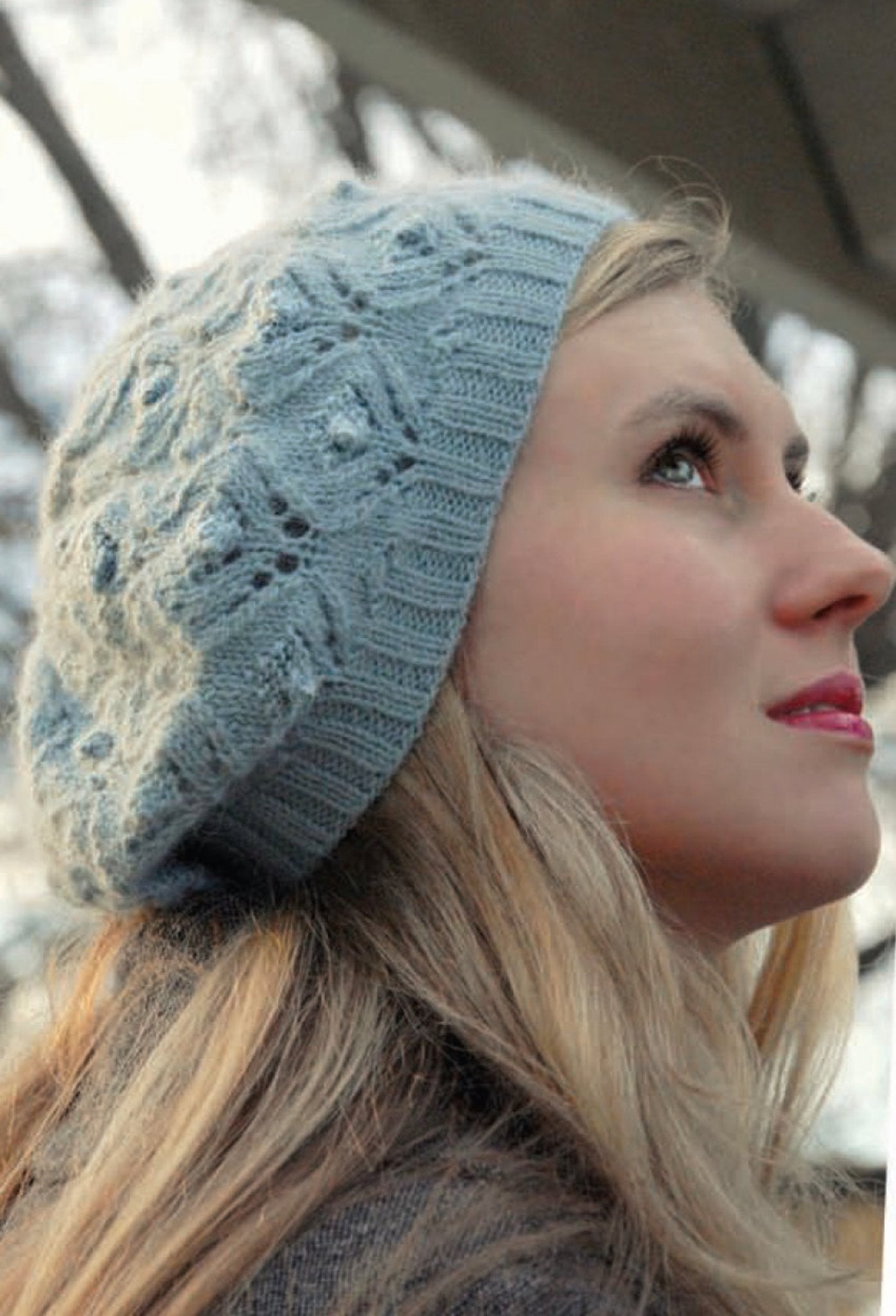 A woman with long blonde hair is gazing upwards. She is wearing the Cloudy Day Beret from Never Not Knitting, featuring an elegant design with lace and bobble stitches in a light blue knit. The background displays out-of-focus tree branches and a glimpse of the sky, hinting at an outdoor environment.